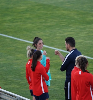 A group of people wearing sports attire are gathered on a grassy field. Some individuals are dressed in red jackets, while others are in turquoise and navy blue. A coach appears to be giving instructions. Sports equipment, such as water bottles and a bench with towels, can be seen nearby.
