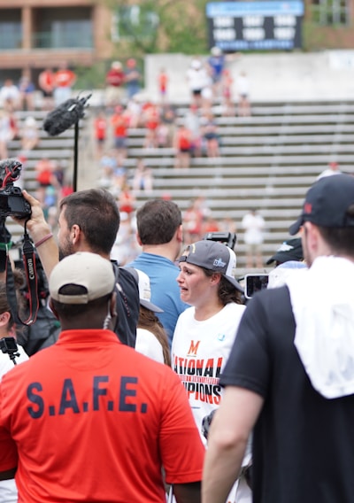 A lively gathering in an outdoor sports venue with a group of people, some wearing matching shirts labeled 'National Champions.' Several individuals are holding cameras and microphones, indicating a media presence. The background shows blurred spectators sitting on bleachers.