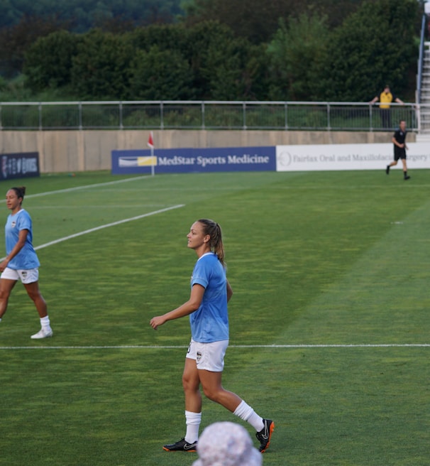 A soccer game scene on a green grass field with players wearing light blue jerseys and white shorts. A goalpost is visible in the background, and there are people sitting or standing on the bleachers. Advertising banners are displayed along the sidelines.