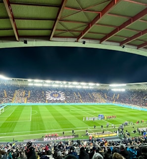 A large football stadium filled with spectators. The field is brightly lit, and two teams are positioned on the pitch. The stadium is covered, with a visible roof structure and stands filled with fans. Scoreboards are visible displaying game information.