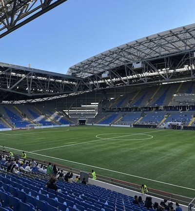 A modern sports stadium with a large, green soccer field and surrounding seating areas. Blue and yellow seats are visible, with some spectators scattered across the stands. The stadium is partially covered with a steel lattice roof, showing a clear blue sky. Several security personnel in high-visibility vests are present.